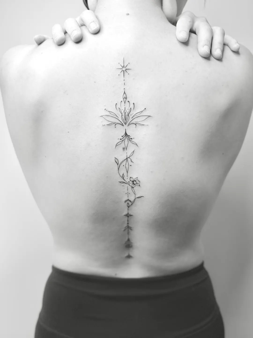 Fine Line Tattoo entlang der Wirbelsäule: Lotusblüte mit zarten Ornamenten und Sternen